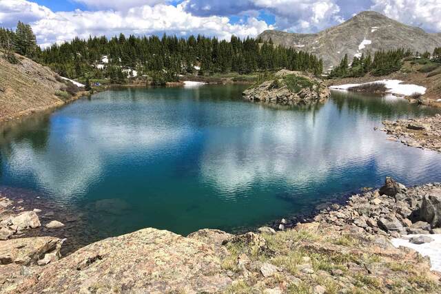 A vivid turquoise alpine lake surrounded by pine trees and rugged mountains under a partly cloudy sky