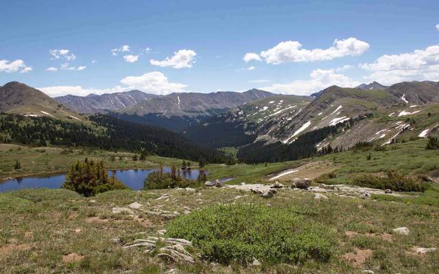 A mountain lake reflecting surrounding peaks and forest, seen from a grassy overlook dotted with boulders