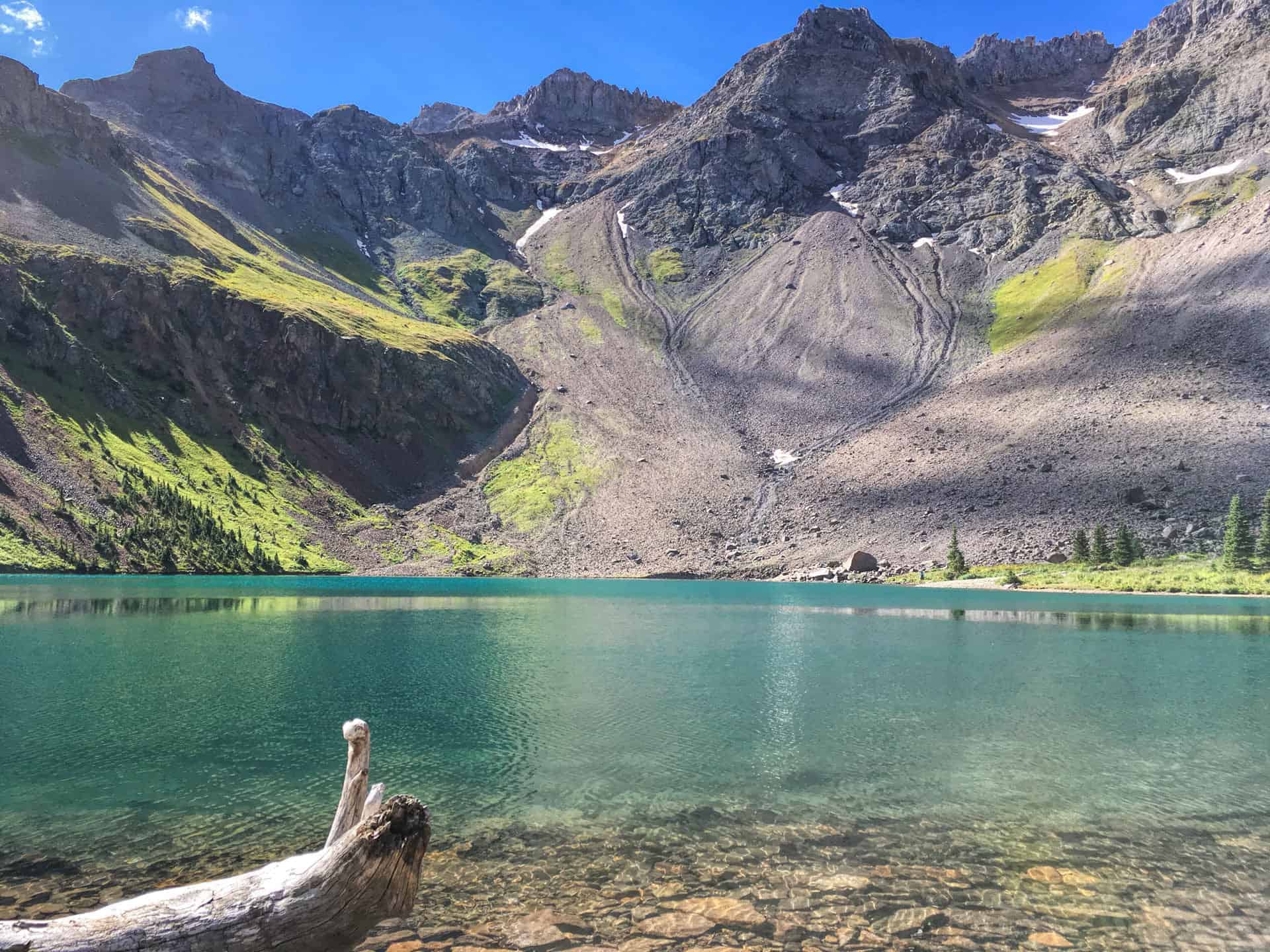 A vivid turquoise alpine lake below a ring of steep, snow-patched peaks and scree slopes, with a bleached log at the water's edge