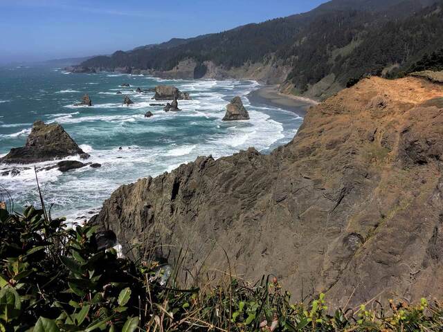 A dramatic view down the rugged Oregon coastline from a high cliff, with sea stacks rising from turquoise surf and forested headlands stretching into the distance