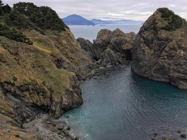 A dramatic view down into a rocky cove with turquoise-blue water, framed by tree-topped cliffs, with a small pebble beach below and misty mountains in the distance